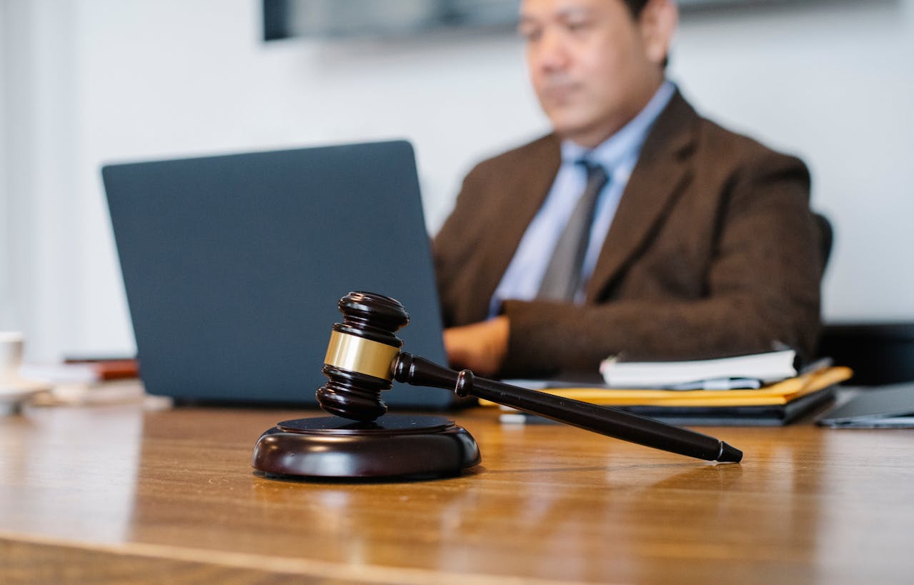 A dark wooden gavel with gold embellishments rests in focus on a light brown desk. In the blurred background is a man working on a laptop. He wears a brown blazer, light blue shirt, and a gray tie.